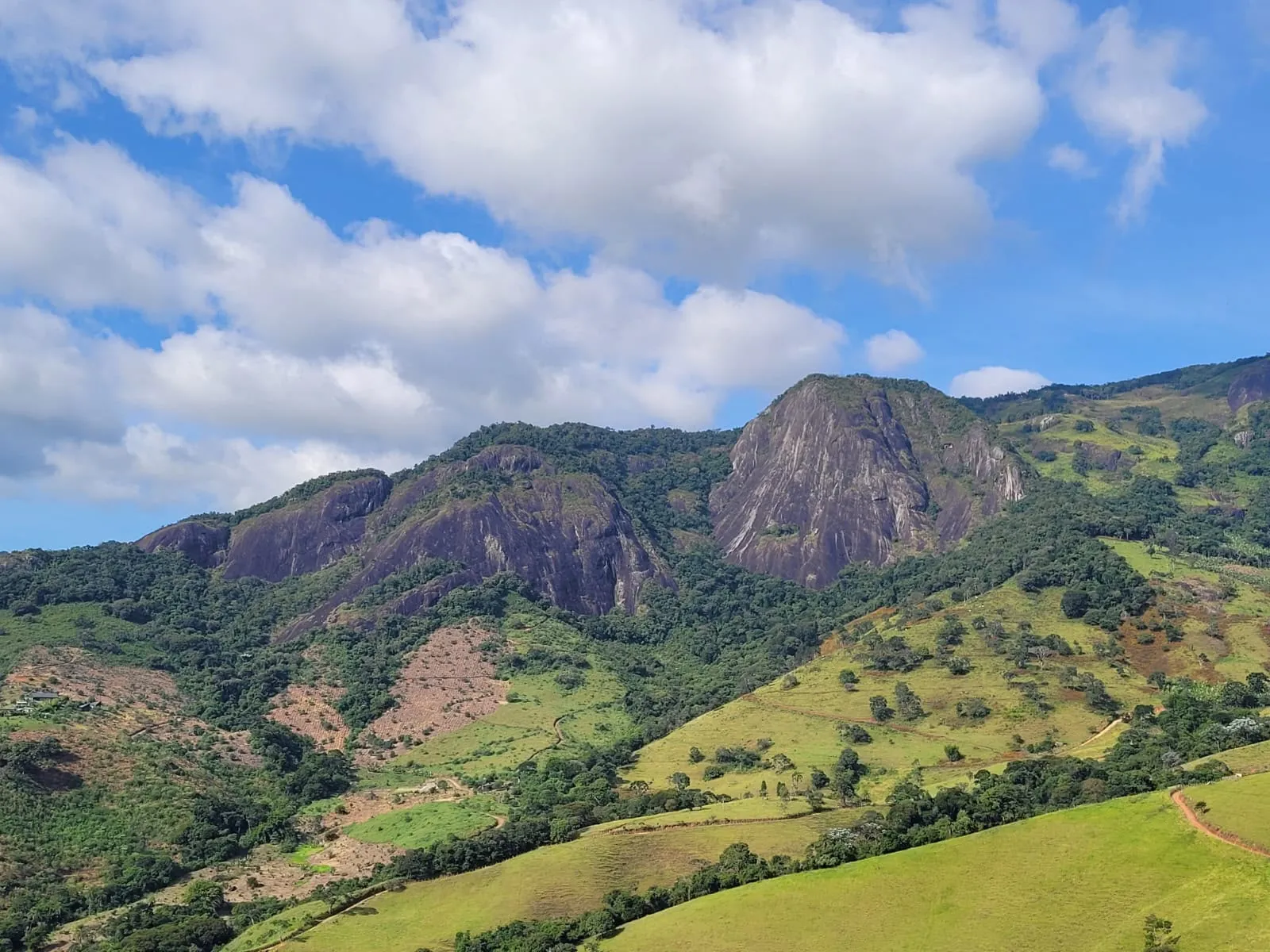 Montanhas de paredões rochosos cercadas por mata e campos verdes sob céu com nuvens