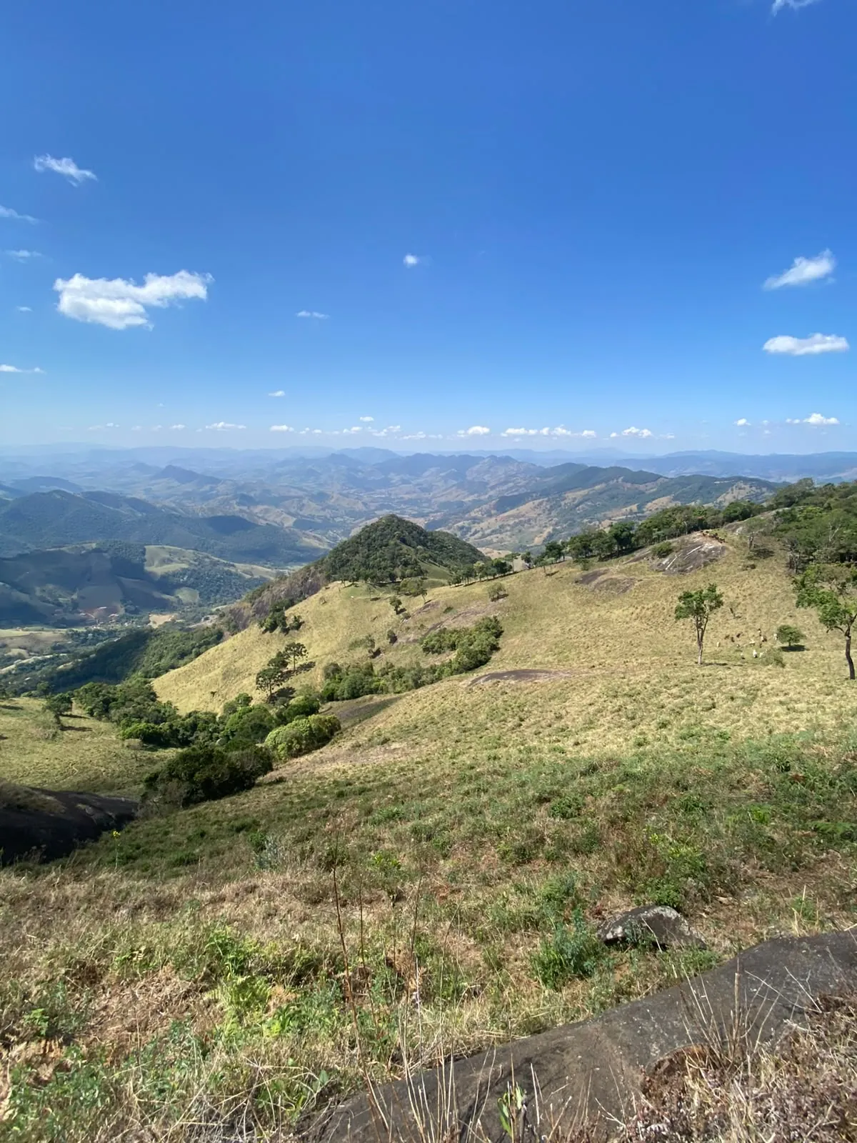 Vista do alto de uma encosta, com campos, árvores e montanhas ao fundo